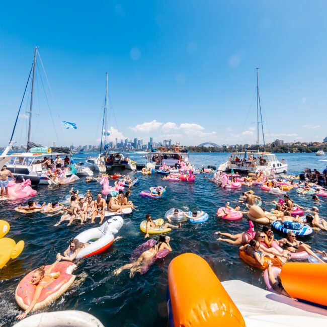 People enjoying a sunny day on the water, surrounded by boats and colorful inflatables. Many are floating and swimming, while others socialize on The Yacht Social Club Sydney Boat Hire boats. The skyline of a city can be seen in the distance, creating a lively and festive atmosphere.