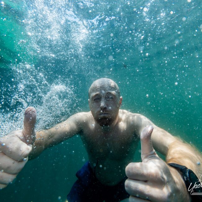 A person underwater gives two thumbs up while surrounded by bubbles. The individual is shirtless and appears content. The scene has an overall greenish-blue tint, typical of underwater environments. A logo in the bottom right corner reads, "The Yacht Social Club Sydney Boat Hire.