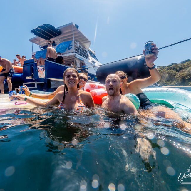 A group of people enjoying a Boat Rental and Parties Sydney The Yacht Social Club event on a yacht. Three individuals are in the water, smiling and holding drinks near a large inflatable float. The yacht in the background has people on board with a "Yacht Social Club" logo visible in the bottom right corner.