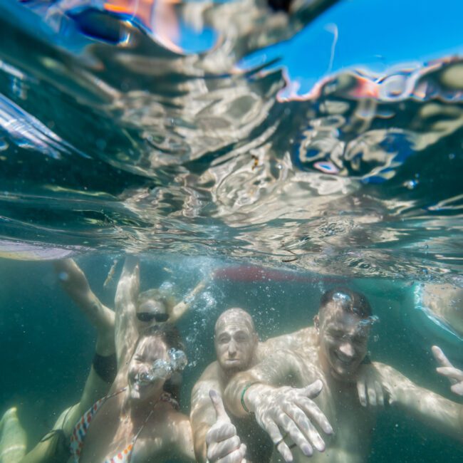 A group of four people is underwater, posing and smiling. One person is wearing a bikini, while the others are in swim trunks. Their hands are extended towards the camera. The water appears clear and blue. The Yacht Social Club Sydney Boat Hire logo is visible in the bottom right corner.