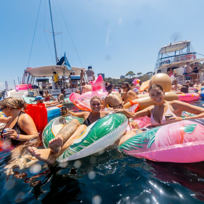 A large group of people enjoying a sunny day on the water, lounging on colorful inflatable floats. They are surrounded by yachts and boats in the background. Some are holding drinks and smiling, creating a festive and lively atmosphere, courtesy of The Yacht Social Club Sydney Boat Hire.