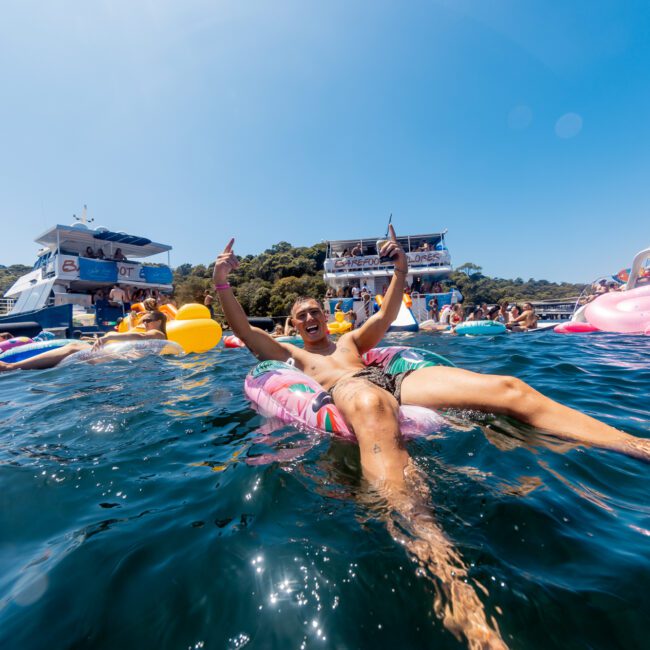 A man floats on an inflatable tube in the ocean, surrounded by other people on various inflatables. Boats and yachts are anchored nearby with people on board, enjoying the sunny day under a clear blue sky. "The Yacht Social Club Event Boat Charters" is written in the bottom right corner.