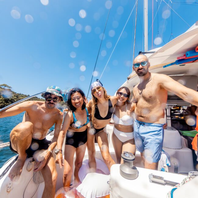 Five people in swimwear smile and pose on a sailboat under a bright, clear sky. The Yacht Social Club event creates a feeling of fun and relaxation among the group. A man is wearing sunglasses and holding a rope on the left, while the lush shoreline and calm blue waters make it perfect for Boat Rental and Parties Sydney.