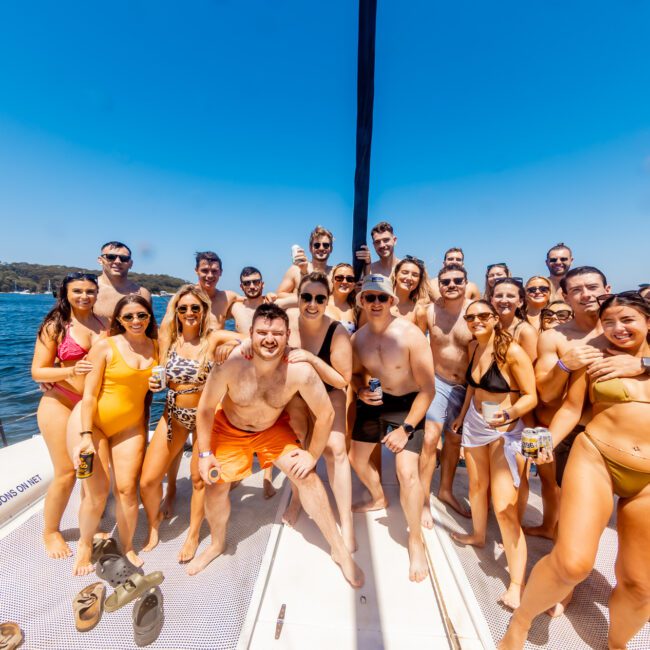 A large group of people in swimsuits poses together on a boat under a clear blue sky. They are smiling and appear to be enjoying a sunny day on the water. The boat is near a shoreline with trees visible in the background. A logo in the corner says "The Yacht Social Club Event Boat Charters.