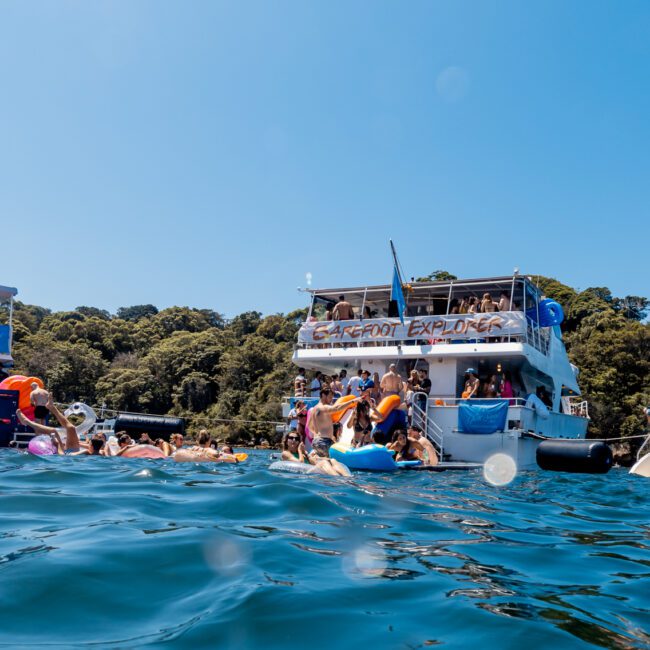 A lively group of people swimming and floating on inflatables near three boats at The Yacht Social Club event. Passengers on the boats enjoy the scenic coastline with dense trees and a clear blue sky, creating a festive atmosphere typical of Sydney Harbour Boat Hire by The Yacht Social Club.