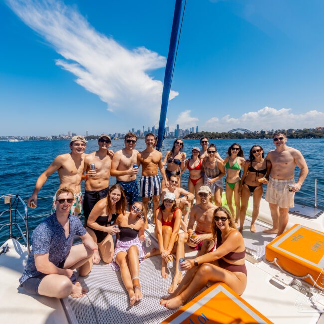 A group of 17 people in swimwear enjoy a sunny day on a sailboat. They are smiling, with some holding drinks, and posing cheerfully with the city skyline and a distinctive bridge visible in the background. The water is blue and the sky is clear during The Yacht Social Club event hosted by Luxury Yacht Rentals Sydney.