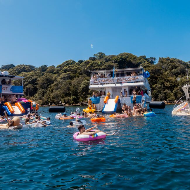 A lively scene with people enjoying a yacht party by The Yacht Social Club. Numerous individuals are swimming and floating on colorful inflatables in the water. Several yachts are docked nearby, and a forested shore is visible in the background. Bright, sunny day with clear blue sky.