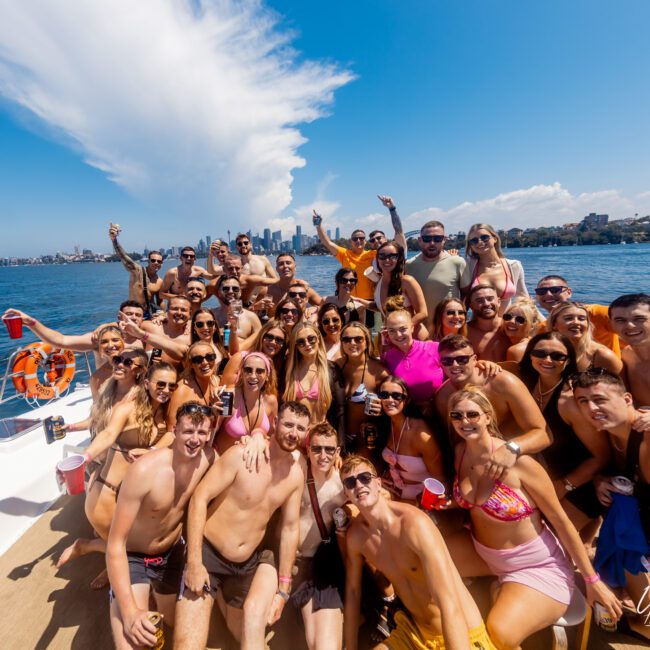 A large group of people, predominantly in swimwear, are gathered on a boat. They are smiling, raising drinks, and posing for the photo against a backdrop of clear blue waters and an expansive skyline. The Yacht Social Club Event Boat Charters ensure a perfect day under the clear, sunny sky with picturesque cloud formations.