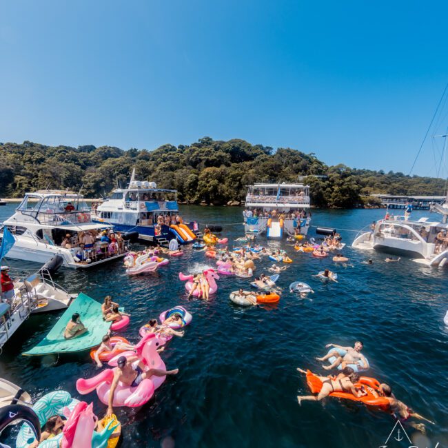 A vibrant scene of a boat party near the coastline, with numerous people floating on colorful inflatables like flamingos, unicorns, and doughnuts. Multiple yachts and party boats from The Yacht Social Club are anchored nearby under a clear blue sky.