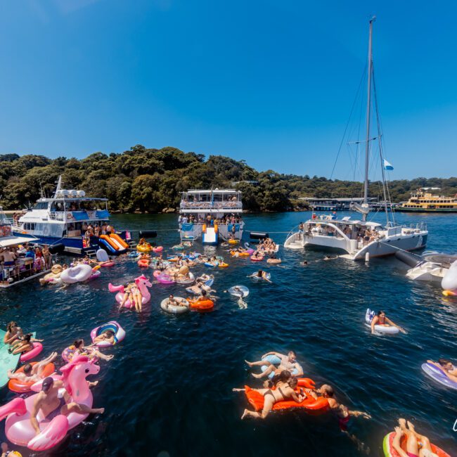 People float on colorful inflatables in the water, surrounded by several anchored boats. The scene takes place on a sunny day with a clear blue sky and lush greenery in the background. The Yacht Social Club Sydney Boat Hire adds to the festive and social atmosphere.