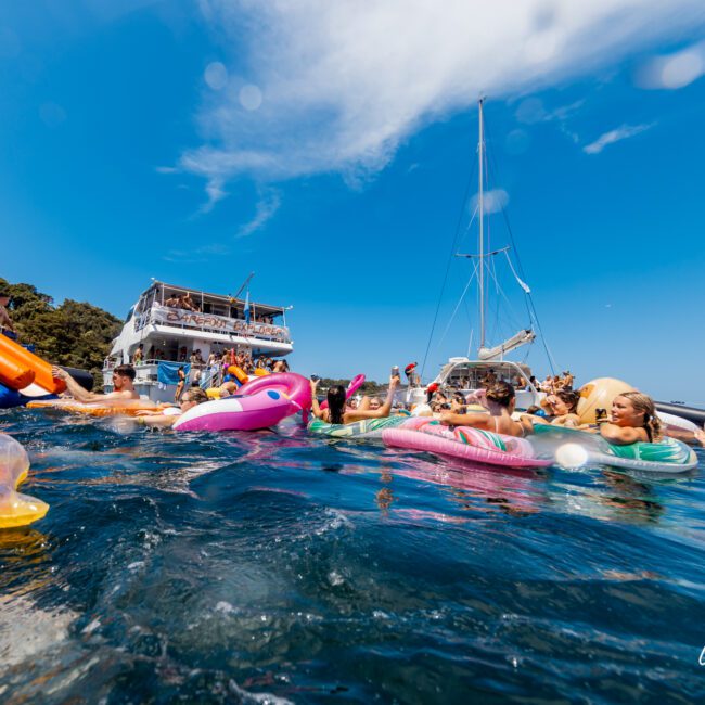 A lively scene of people enjoying a sunny day on the water. Various individuals float on colorful inflatables, including unicorns and tubes, near two anchored boats. The sky is clear blue, and the event logo "The Yacht Social Club" is visible in the bottom right corner, promoting Boat Parties Sydney The Yacht Social Club.