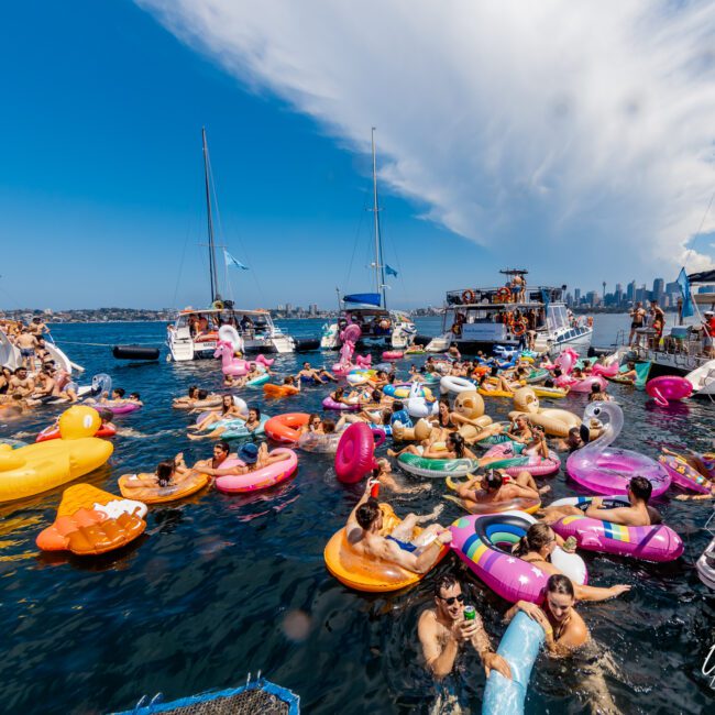 A lively scene of people enjoying a sunny day on the water, floating on colorful inflatable devices near anchored boats. The sky is clear with a few clouds and the distant city skyline is visible. The atmosphere is festive and relaxed as if it were part of The Yacht Social Club Event Boat Charters in Sydney.