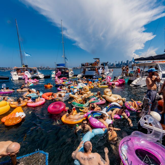 A lively scene of a crowded boat party on the water featuring numerous people enjoying the sun on colorful inflatable rafts. There are several boats in the background against a bright, clear sky with some clouds. The atmosphere is festive and cheerful, epitomizing Boat Parties Sydney The Yacht Social Club.