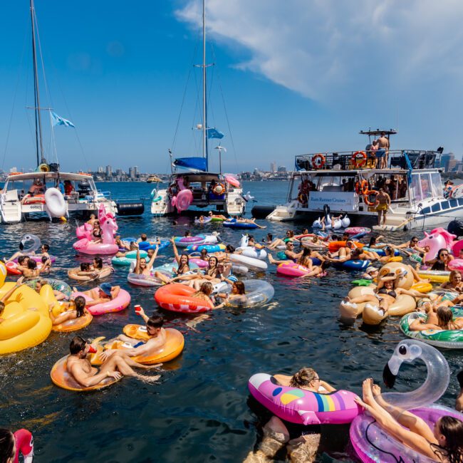 A lively pool party scene at The Yacht Social Club Sydney Boat Hire shows numerous people in colorful inflatable floats of various shapes and sizes enjoying the water between anchored boats. Some people are dancing on the boats, and the sunny sky contrasts with the city skyline in the background.