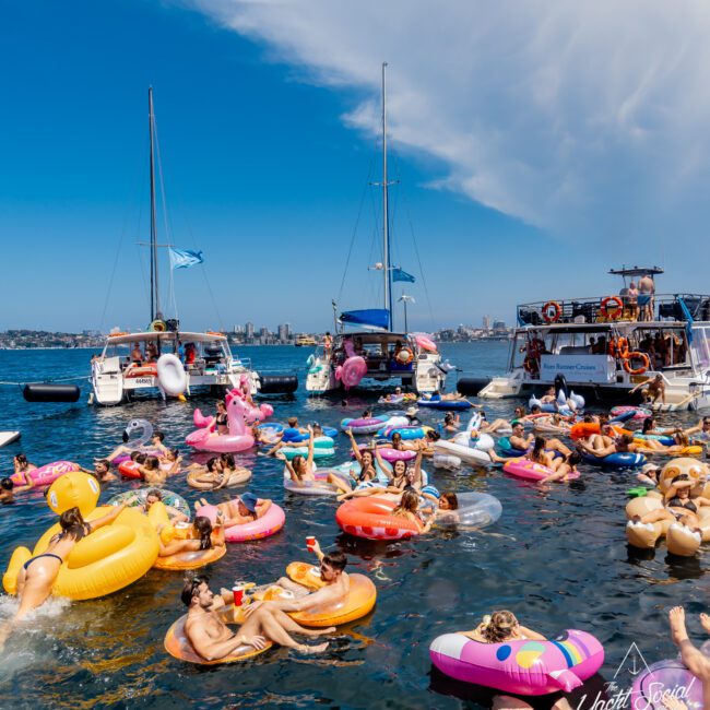 A lively scene of people enjoying a sunny day on the water, floating on colorful inflatable rafts and tubes. Multiple boats from The Yacht Social Club are docked in the background under a clear sky. Some individuals are holding drinks and socializing, adding to the festive atmosphere created by The Yacht Social Club Sydney Boat Hire.