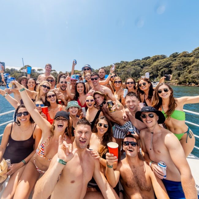 A large group of people in swimwear are gathered on a boat, smiling and holding drinks. They appear to be enjoying a sunny day on the water with trees and buildings visible in the background. Everyone is wearing sunglasses, some making celebratory gestures—perfect vibes for The Yacht Social Club Sydney Boat Hire.