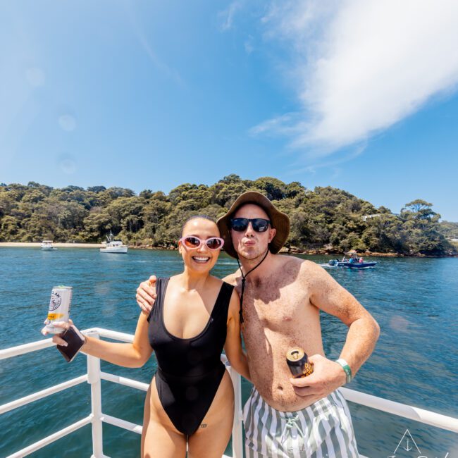 A woman in a black swimsuit and sunglasses and a man in striped shorts and a sun hat, both holding beverages, stand on a boat from The Yacht Social Club with their arms around each other. A tree-covered island and other boats are visible in the background on a sunny day.