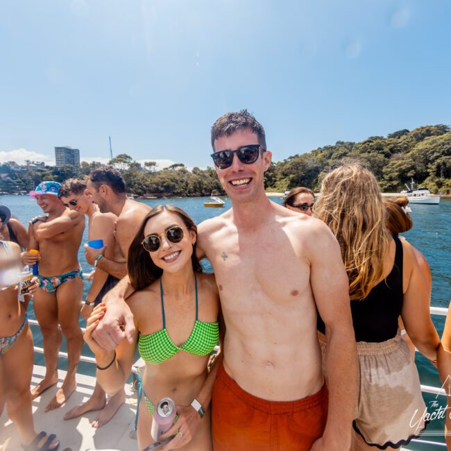 A group of people wearing swimsuits enjoy a sunny day on a boat. In the foreground, a woman in a green bikini and sunglasses stands next to a man in red swim trunks and sunglasses. Others in the background are socializing, with water and trees visible behind them—a typical scene at The Yacht Social Club Event Boat Charters.