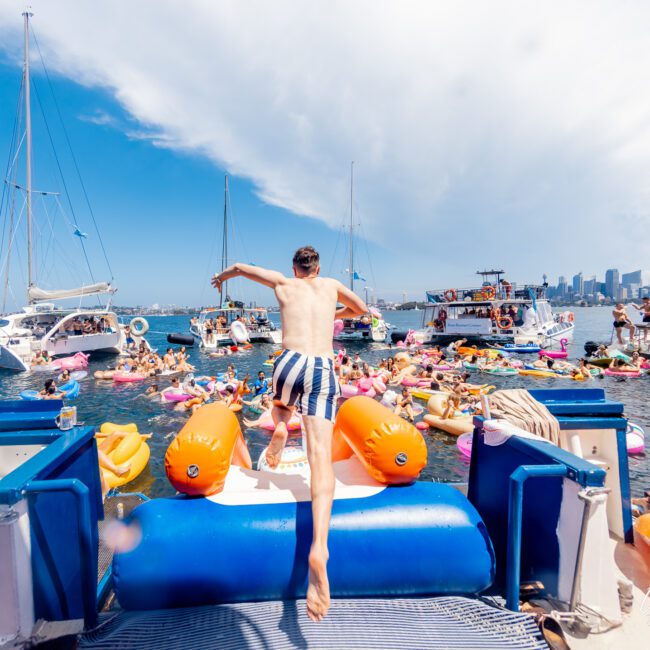 A person jumps off a boat rental into a crowded water area filled with colorful inflatables and people enjoying the summer. Sailboats are anchored nearby, and a city skyline is visible in the background under a partly cloudy sky, adding to the festive flair of The Yacht Social Club Event.