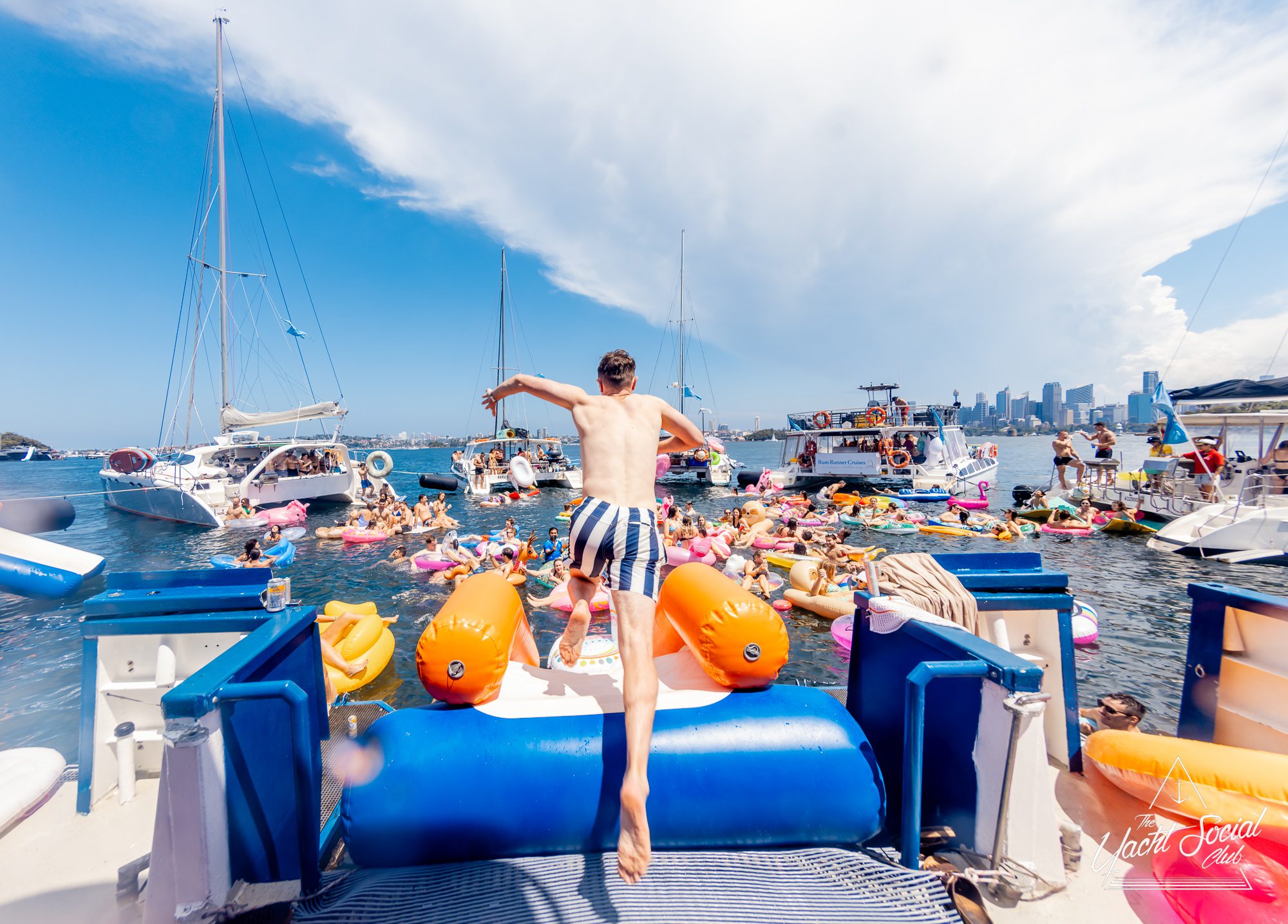 A person jumps off a boat rental into a crowded water area filled with colorful inflatables and people enjoying the summer. Sailboats are anchored nearby, and a city skyline is visible in the background under a partly cloudy sky, adding to the festive flair of The Yacht Social Club Event.