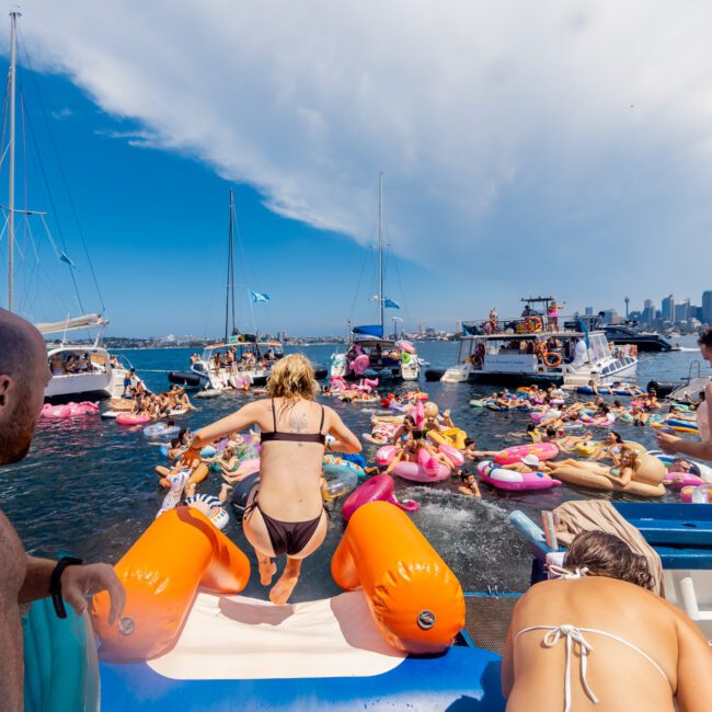 A lively scene of people enjoying a sunny day on the water. Some are jumping off a boat into the sea, while others float on inflatable pool toys nearby. Several boats from The Yacht Social Club Sydney Boat Hire form a backdrop, and the sky is mostly clear with a few clouds in the distance.