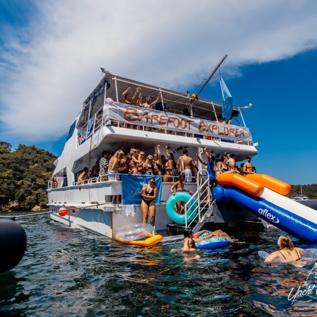 People are enjoying a sunny day on a large double-decker party boat named "Barefoot Explorer" at The Yacht Social Club. Some are on the boat, while others swim, float, or use a large inflatable slide into the clear water. The boat is anchored near a forested shoreline.