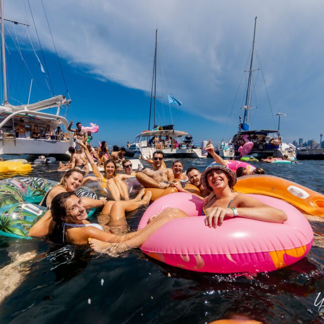 A group of people is enjoying a sunny day on the water, lounging on colorful inflatable floats, surrounded by boats. They are smiling, waving, and appear to be having a great time at The Yacht Social Club Event Boat Charters. The sky is clear with a few scattered clouds.