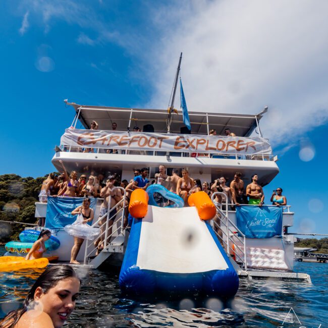 A large boat named "Barefoot Explorer" is anchored with many people on board, some sitting and some standing. It features a water slide with several people in the water nearby. Blue skies and calm waters create a lively and sunny atmosphere, ideal for The Yacht Social Club Sydney Boat Hire experience.