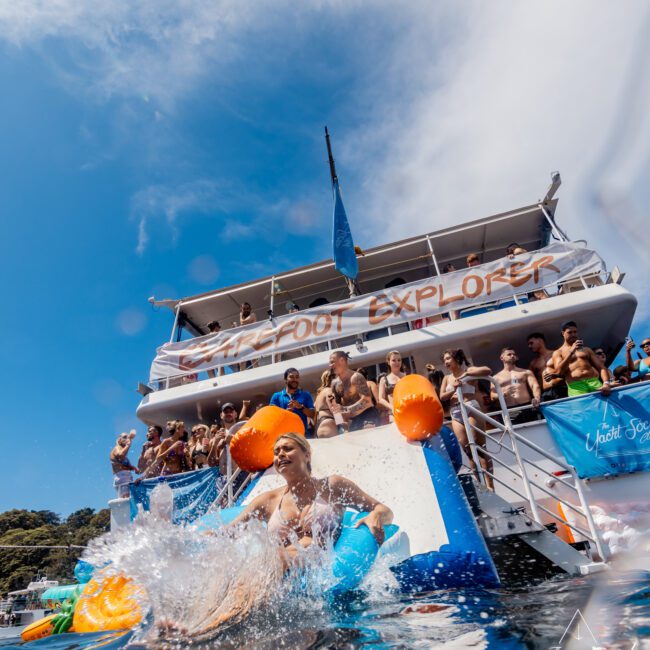 A person splashes into the water from a large boat named "Sunkissed Explorer" filled with people, enjoying The Yacht Social Club Sydney Boat Hire. The boat has blue and orange floats, and the clear sky adds to the lively, recreational atmosphere. Other guests watch and relish the moment.