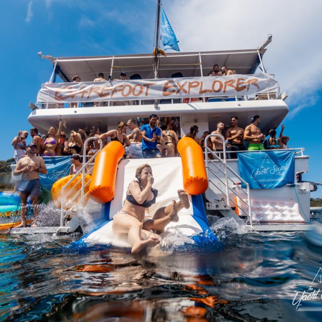 People enjoy a sunny day on a boat named "Barefoot Explorer" with many swimming and some using an inflatable slide into the water. A mix of bright swimwear and water toys creates a lively scene under a clear sky, much like the events hosted by The Yacht Social Club.