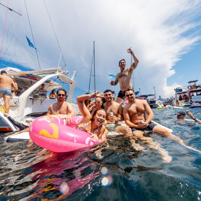 A group of six friends in swimsuits enjoying a lively day at sea, sitting on and around a large pink flamingo float near several anchored boats. The friends are smiling, some holding drinks, with a clear blue sky and a few clouds in the background—an ideal scene for The Yacht Social Club Boat Parties Sydney.