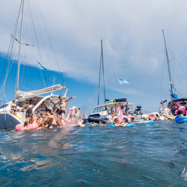 A lively scene of people enjoying a sunny day on the water with sailboats and inflatables. Various individuals are relaxing and swimming near the boats, with colorful floats shaped like unicorns and flamingos adding to the festive atmosphere. The Yacht Social Club logo, known for Luxury Yacht Rentals Sydney, is visible.