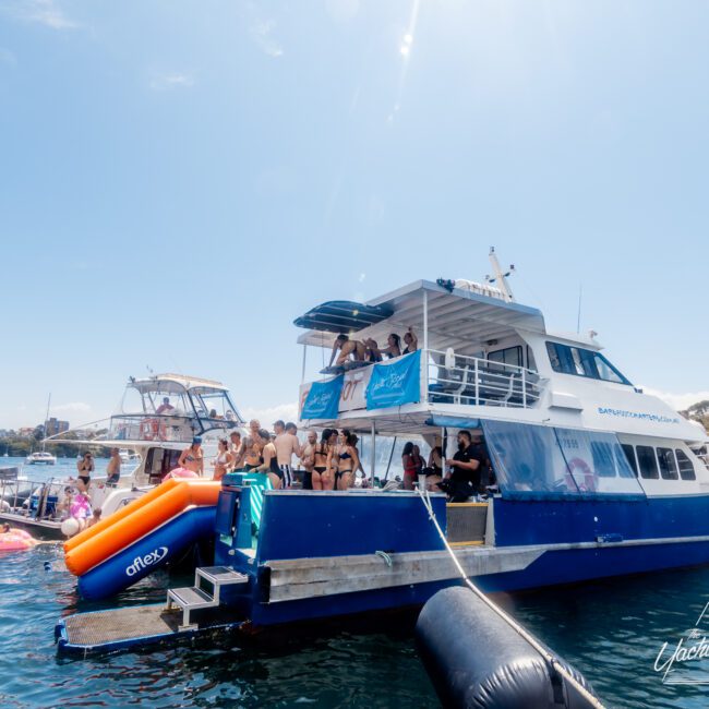 A group of people enjoying a sunny day on a large blue and white yacht. Some are on the upper deck, while others are socializing on the main deck. An inflatable slide extends from the yacht into the water. Several smaller boats are nearby, capturing the essence of a Boat Party with The Yacht Social Club in Sydney Harbour.