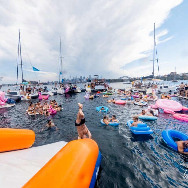 A lively scene on the water with numerous people enjoying themselves on brightly colored inflatables. Various boats from The Yacht Social Club Sydney Boat Hire surround the group, and a city skyline with tall buildings can be seen in the background under a partly cloudy sky.