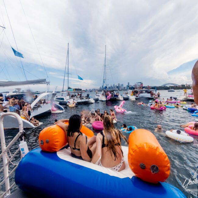 A lively group of people enjoy a day on the water, floating on various inflatables near anchored yachts and boats. The scene is festive with participants in swimsuits surrounded by colorful floats. The sky is partly cloudy, and a city skyline is visible in the background—a perfect setting for The Yacht Social Club Event Boat Charters.