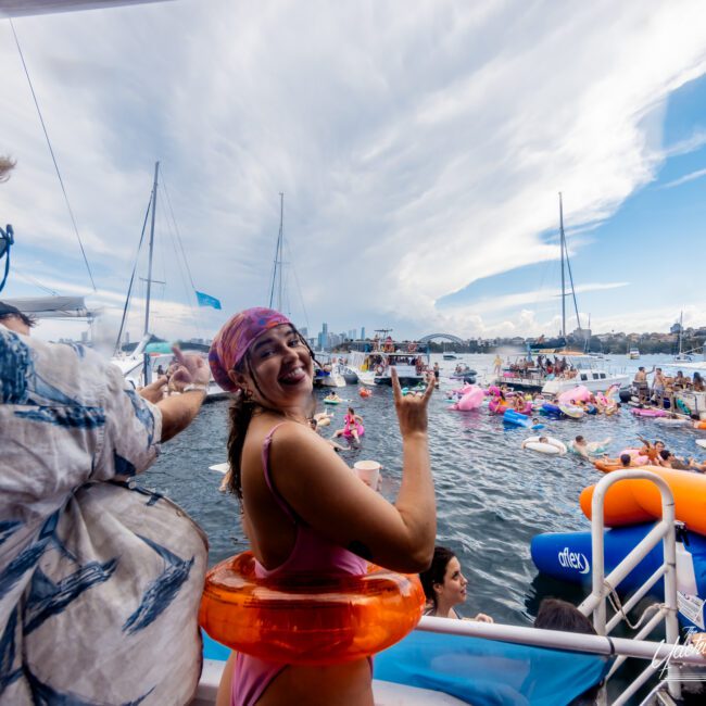 A smiling person in a pink swimsuit and swim ring gestures towards the camera while standing on a boat. In the background, many people are enjoying the water with colorful inflatables. Sailboats and a cloudy sky can be seen. The Yacht Social Club Sydney Boat Hire creates a festive and lively atmosphere.