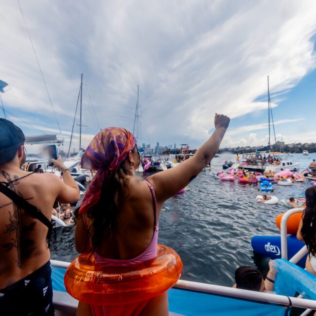 People in swimwear on a boat from The Yacht Social Club raise their arms and cheer while looking towards a crowd in the water with various inflatables. Sailboats are in the background under a dramatic sky. The atmosphere is festive and lively, capturing the essence of Sydney Harbour Boat Hire.