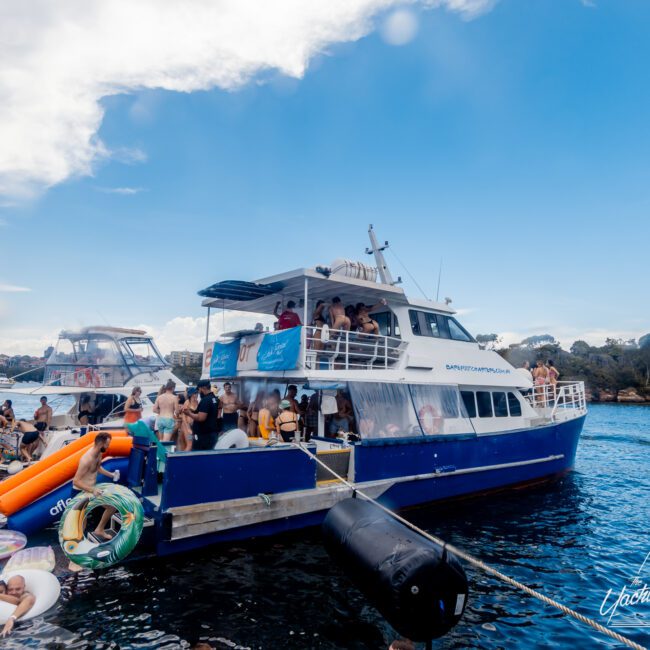 A large blue and white party boat is filled with people socializing and enjoying a sunny day on the water, organized by Sydney Harbour Boat Hire The Yacht Social Club. Some people are on inflatables in the water. Another boat is nearby, and land can be seen in the background. The sky is mostly clear with some clouds.