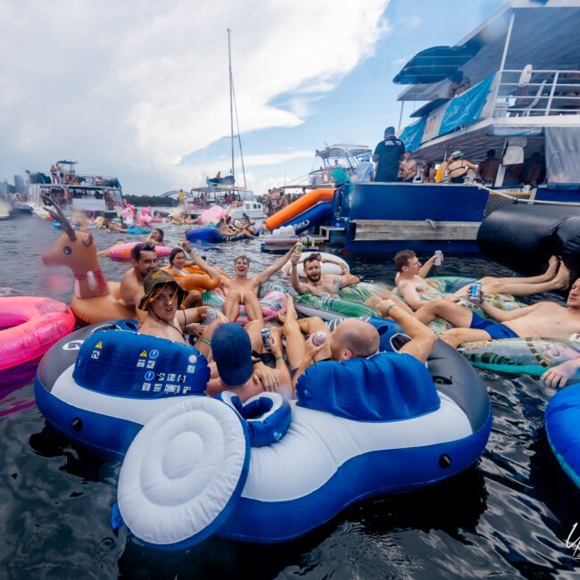 A group of people relax on inflatable rafts and floaties in the water, surrounded by yachts and boats from The Yacht Social Club Sydney Boat Hire. They appear to be enjoying a social gathering under a partly cloudy sky. The scene conveys a lively and festive atmosphere.