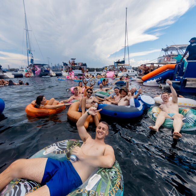 A group of people floating on inflatable tubes and floats in a large body of water, smiling and raising their hands. Various boats are anchored nearby, and the sky is partly cloudy. A logo at the bottom right reads "The Yacht Social Club Sydney Boat Hire.