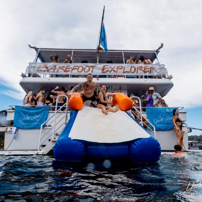 A lively scene on the luxury yacht "Barefoot Explorer." People are enjoying various activities, with one person sliding into the water on a large inflatable slide. The boat is anchored in a scenic location with clear water and people swimming nearby, epitomizing Boat Parties Sydney The Yacht Social Club.