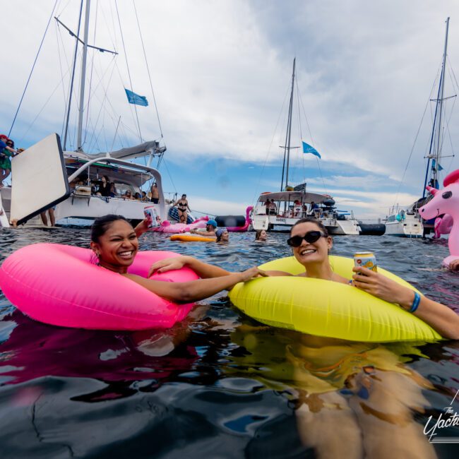 Two people are floating in the water with inflatable rings, one pink and one yellow. They are smiling and holding hands. Sailboats and a luxury yacht from The Yacht Social Club Event Boat Charters are visible in the background along with other people enjoying the festive atmosphere.