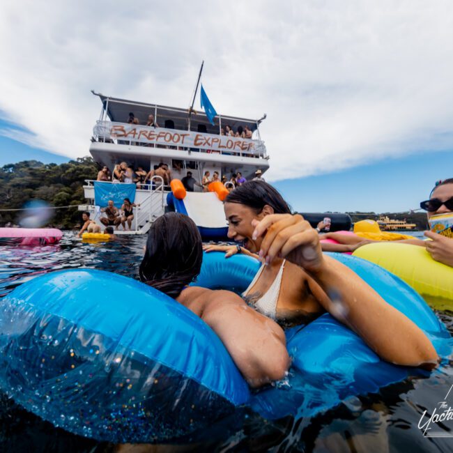 A group of people enjoy a day on a lake, floating on inflatable rafts. In the background, a large boat named "Barefoot Explorer" is anchored, with others on board and using a slide that descends to the water. The scene is lively and sunny—a perfect day reminiscent of Boat Parties Sydney The Yacht Social Club.