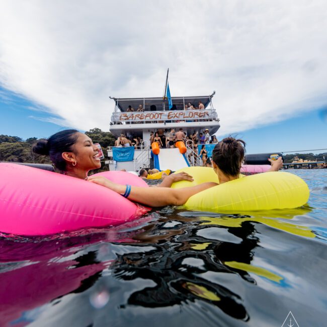 Two people relaxing on colorful inflatable rings in the water near a boat named "Barefoot Explorer." The boat, part of The Yacht Social Club Event Boat Charters, has several people onboard and a blue flag is flying. The sky is partly cloudy, and the setting appears to be a recreational boating area.