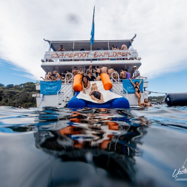 A group of people relaxes on and around a boat named "Barefoot Explorer." Several individuals are on an inflatable floating platform in the water. The scene is lively, with people enjoying the sunny day and scenic backdrop of water and greenery—perfect for a Yacht Social Club Sydney Boat Hire experience.