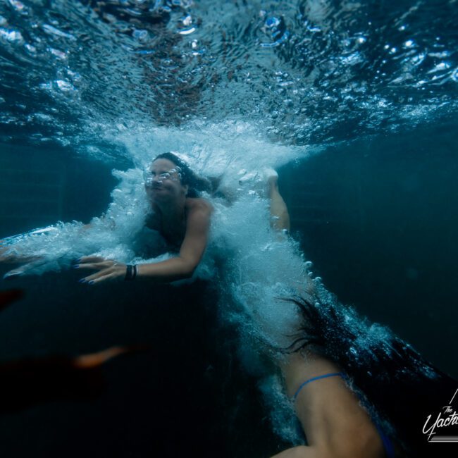 Two people are swimming underwater in a pool, creating bubbles and waves. The person on the left extends their arms forward while looking ahead, and the person on the right is partially submerged, with long hair flowing behind them. The logo "The Yacht Social Club Sydney Boat Hire" is visible in the corner.