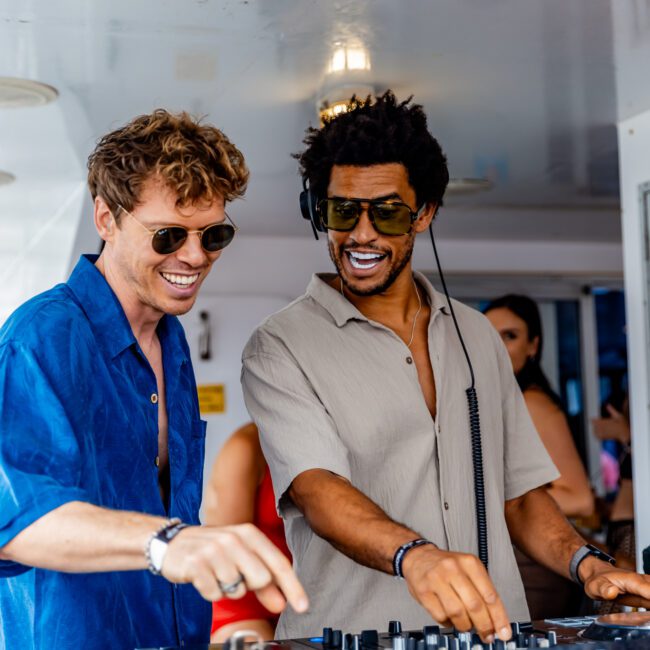 Two men are DJing on a yacht, smiling and enjoying the moment. One wears a blue shirt and sunglasses, while the other wears a beige shirt and sunglasses. Both are adjusting music equipment, with people and a bright, airy setting in the background at The Yacht Social Club Sydney Boat Hire.