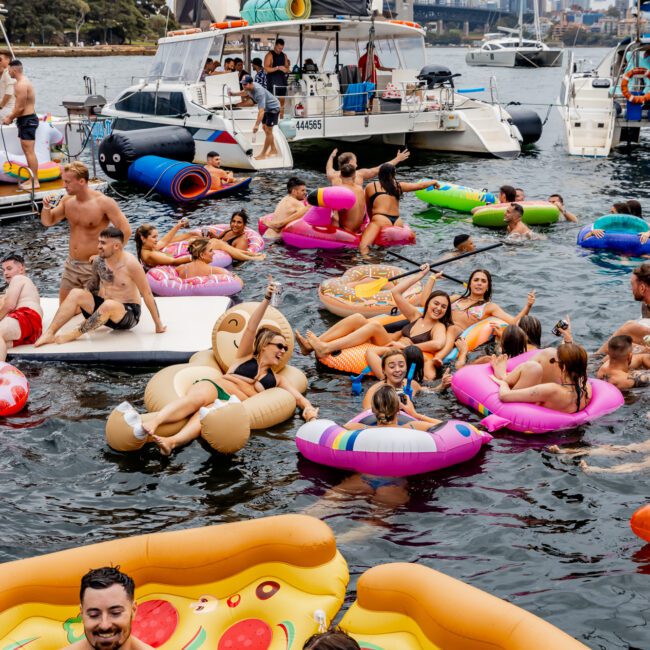 A lively group of people enjoying a pool party on various inflatable floaties, including a giant pizza slice, donuts, and a teddy. Multiple boats are anchored nearby as part of The Yacht Social Club Event Boat Charters, with a prominent bridge and city skyline visible in the background under a cloudy sky.