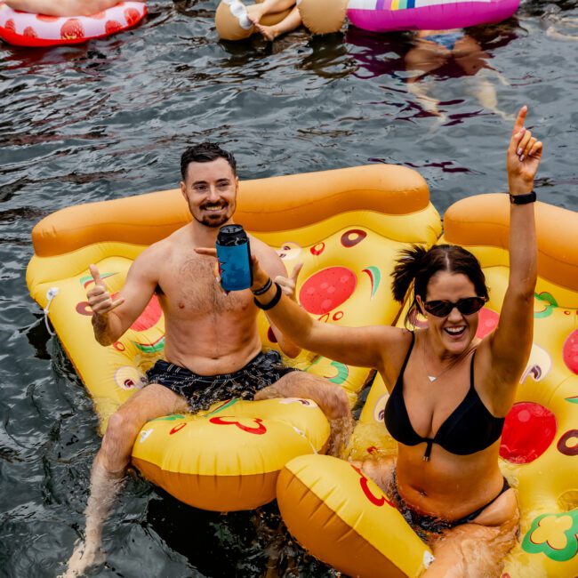 A festive scene of people enjoying a pool party. In the foreground, a man and woman in swimsuits are lounging on a pizza-shaped inflatable float. The man is holding a phone, while the woman is raising her arm and smiling. Other partygoers float nearby, evoking vibes reminiscent of luxury yacht rentals in Sydney Harbour.