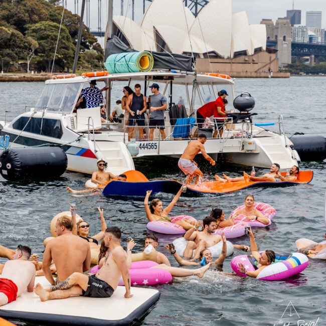 A group of people enjoys a vibrant party on inflatable floats in the water near a docked luxury yacht from Boat Rental and Parties Sydney The Yacht Social Club. The Sydney Opera House and Harbour Bridge are visible in the background. Some people are on the boat, while others relax in the water holding drinks.